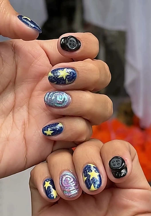 Dos manos con uñas cortas pintadas a mano: estrellas amarillas sobre azul con glitter, espirales tornasol y botones negros
