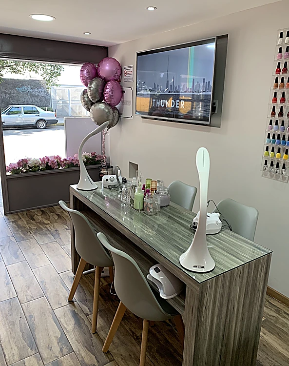 Mesa de manicure con lámparas blancas, sillas gris claro, globos rosas y un ventanal con jardinera de flores