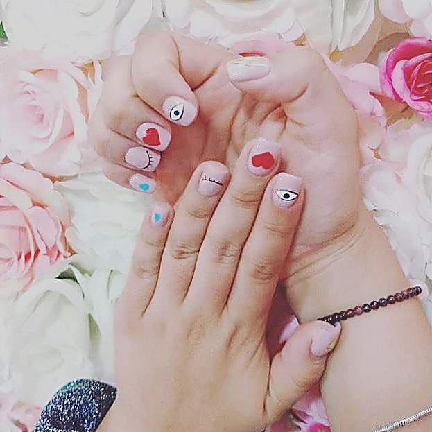 Uñas cortas nude con corazones rojos, ojitos y puntos azules, frente al muro de rosas