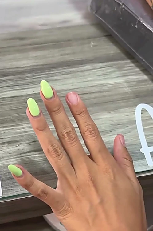 Mano sobre la mesa de manicure con uñas almendradas, unas en verde neón y otras en rosa nude